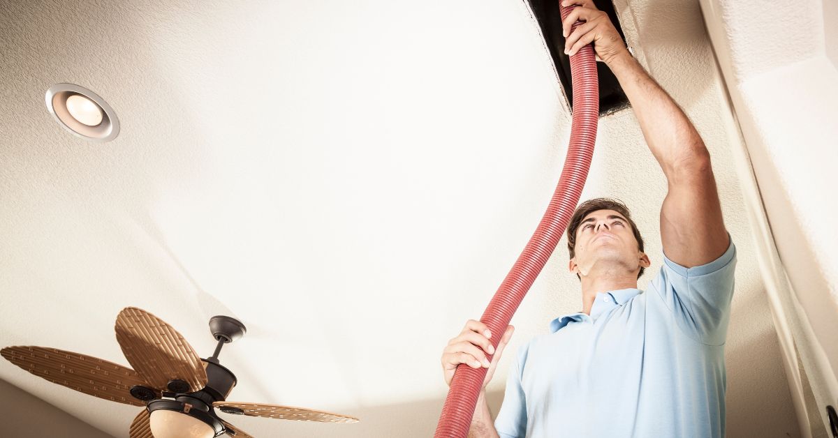 A person in a blue shirt uses a large red hose to perform air duct cleaning on an air duct in the ceiling. A ceiling fan and recessed light are also visible.