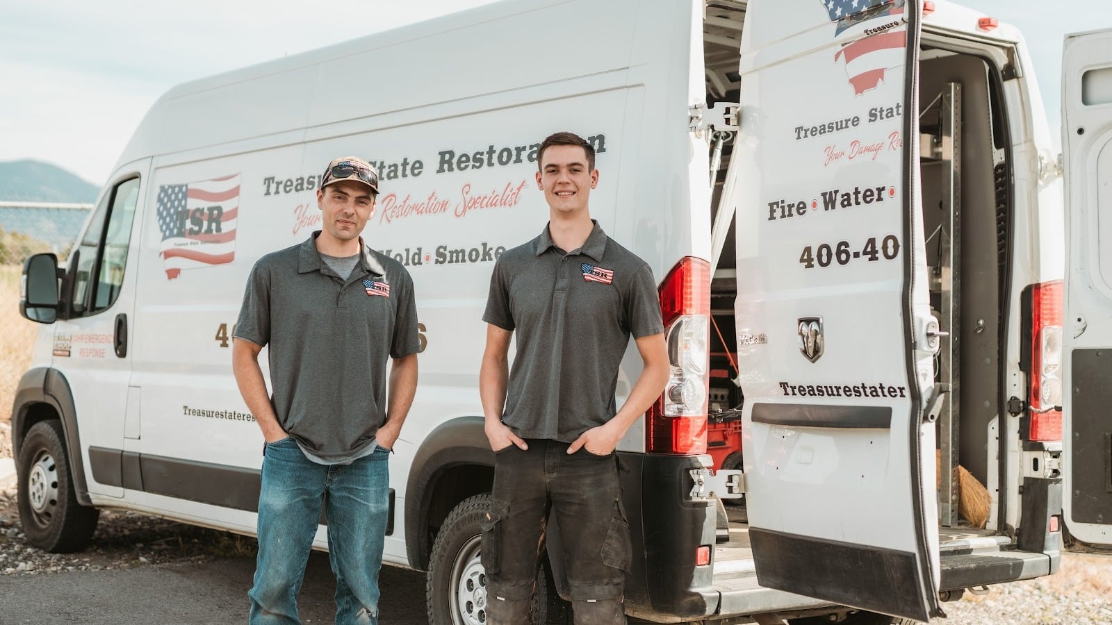 Two workers in uniform stand in front of a white Treasure State Restoration van with doors open, posing and smiling outdoors, ready to provide emergency response services for any commercial property damage.
