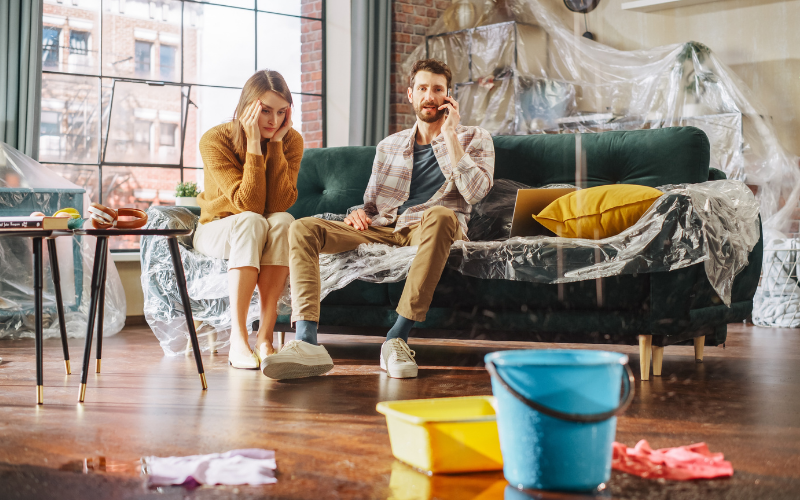 A man and woman sit on a sofa in a living room with water leaking onto the floor; buckets catch the water, plastic covers some furniture, and they consider calling Restoration Services for help.