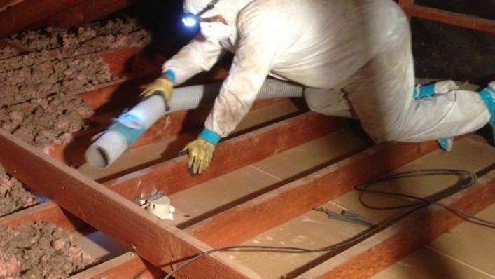 Person in protective gear with headlamp vacuuming insulation from an attic between wooden floor joists, as part of an emergency response to potential damage in a commercial property.