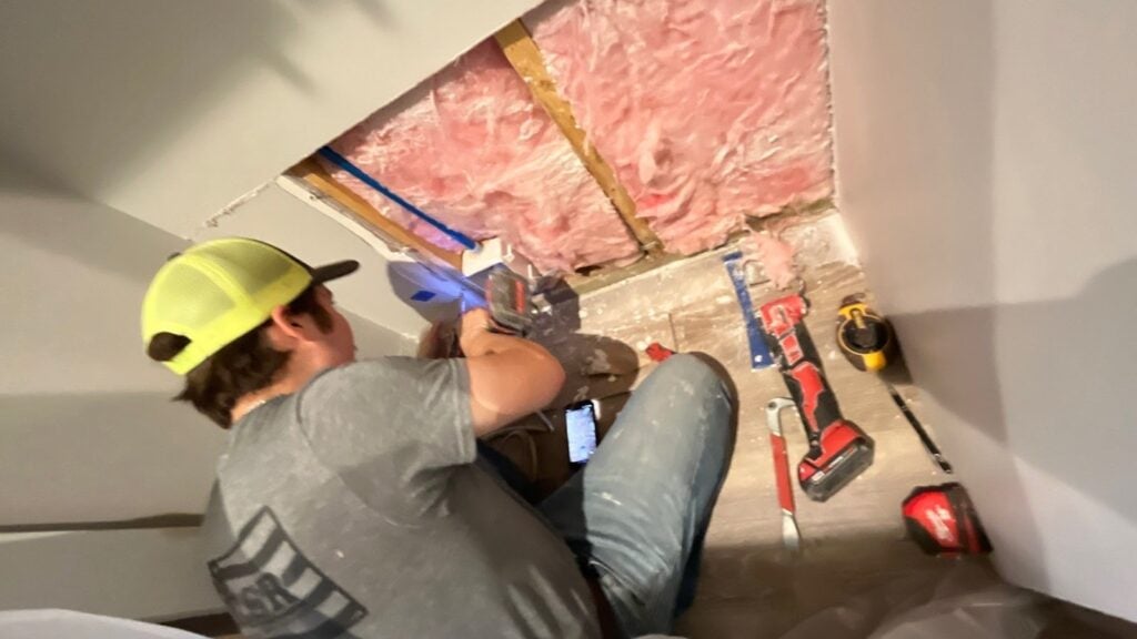 A person wearing a yellow cap is using a power tool to work on exposed insulation and pipes in a wall, illustrating the critical emergency response often required within the first 24 hours after commercial property damage. Various tools are scattered on the floor nearby.