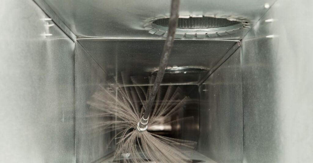 A cylindrical brush attached to a rod is cleaning the inside of a metal air duct, demonstrating effective air duct cleaning techniques.