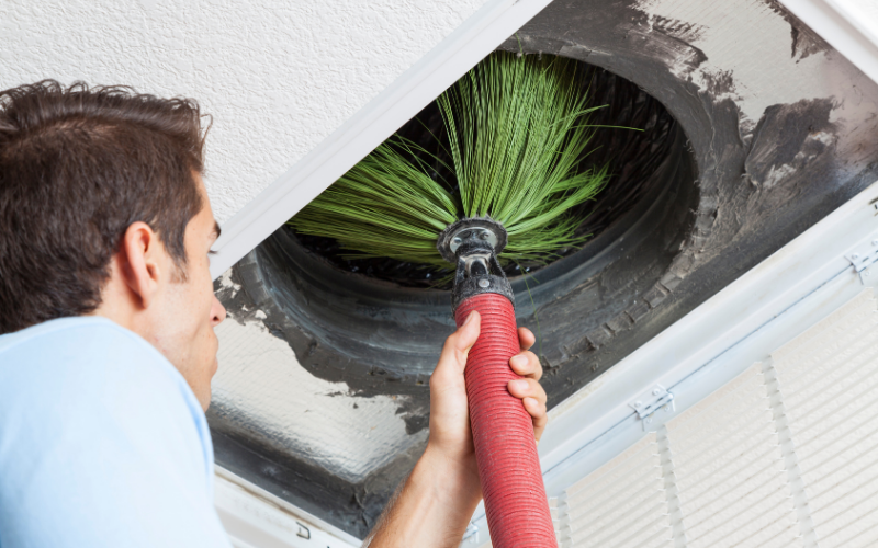 A person uses a brush attached to a hose to clean the inside of an air duct mounted in a ceiling.
