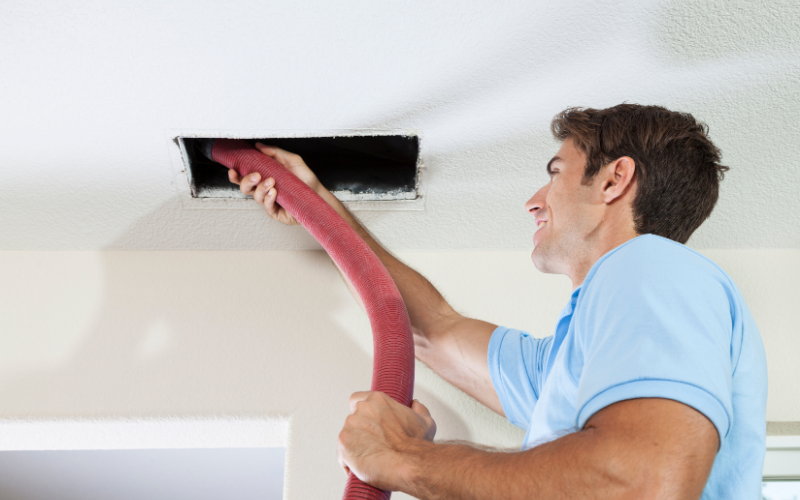 Man performing air duct cleaning with a vacuum hose on a ceiling vent in a residential home.