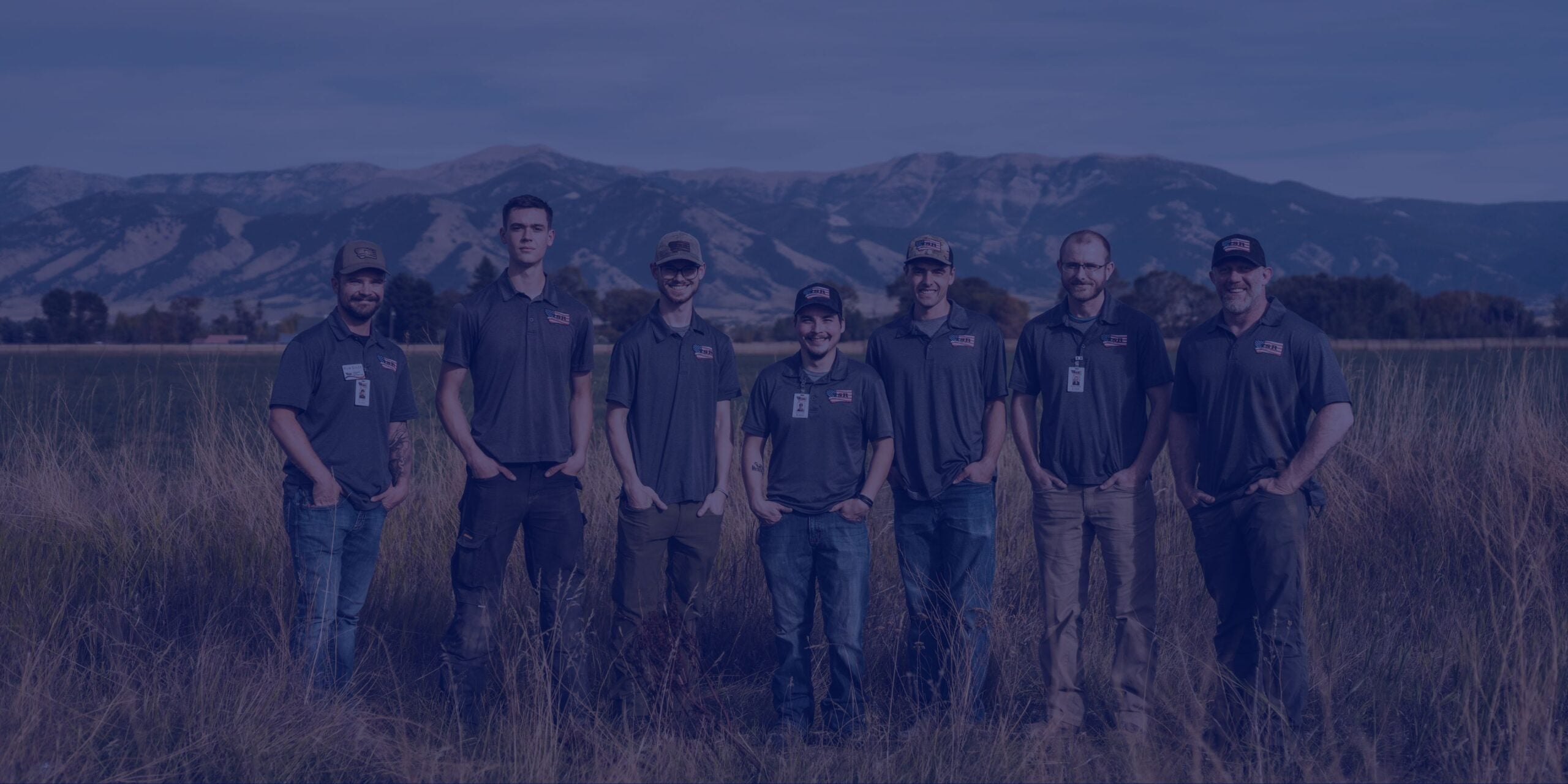 Eight men wearing matching shirts and ID badges stand in a field with mountains in the background, posing for a group photo as part of their restoration services team.