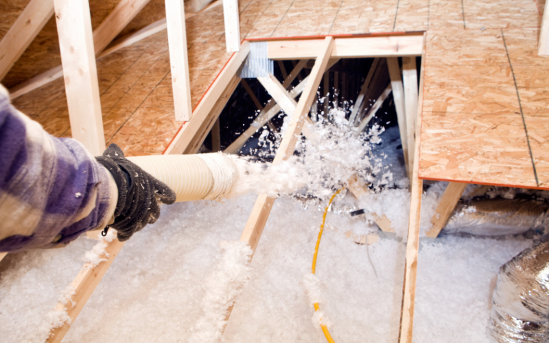 A person sprays loose-fill insulation into an unfinished attic between wooden floor joists after completing insulation removal.