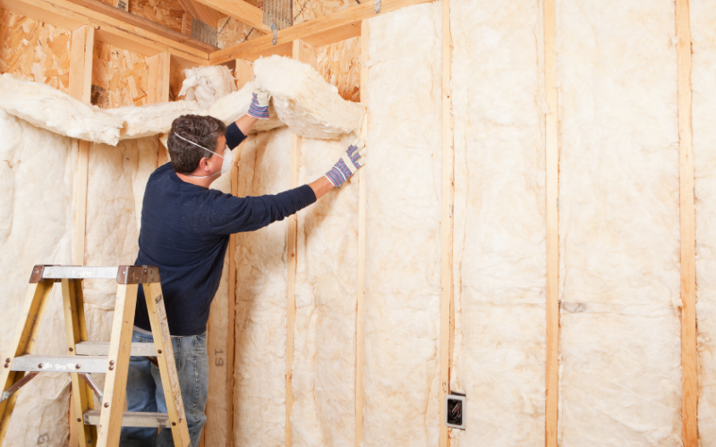A person standing on a ladder installs fiberglass insulation in an unfinished interior wall of a house under construction, demonstrating professional insulation services that ensure energy efficiency and comfort throughout the home.