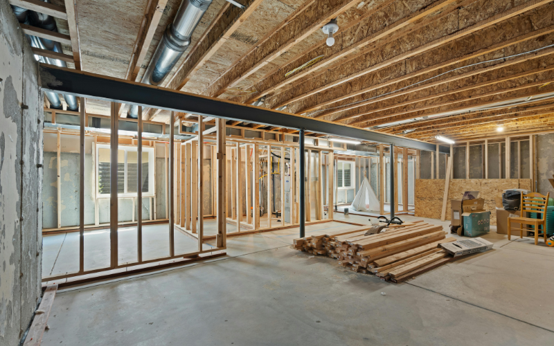 Unfinished basement with exposed wooden framing, steel beam, visible ductwork, concrete floor, window, and construction materials stacked on the ground—ideal space for insulation removal or other insulation services to help prepare your home for future renovations.