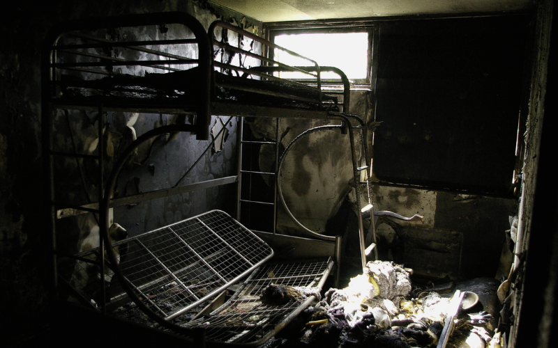 A burned and damaged bunk bed stands in a dark, charred room with debris and singed belongings scattered on the floor, awaiting professional Restoration Services to return the space to its former condition.