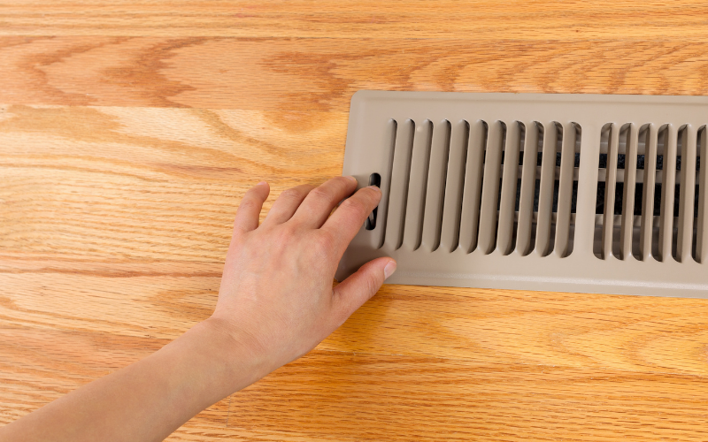A hand adjusts the lever on a floor air vent set in a wooden floor, showcasing the importance of regular air vent cleaning to maintain optimal airflow and indoor air quality.