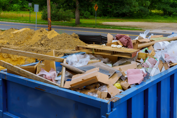 A blue dumpster filled with construction debris, cardboard, wood, and plastic sits near a roadside with trees and dirt piles in the background—evidence of ongoing repair and restoration services transforming the area.