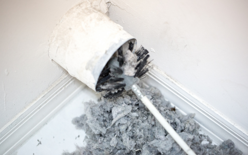 A lint brush is cleaning excess lint from a dryer vent in the corner of a room, with a pile of gray lint on the floor—a simple step that highlights the importance of regular air vent cleaning to maintain safety and efficiency.