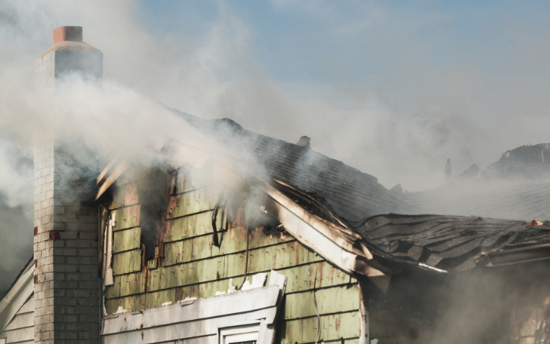 Smoke rises from the damaged roof of a house with green siding and a brick chimney, indicating recent fire damage—a situation where skilled restoration professionals are essential to repair and restore the property.