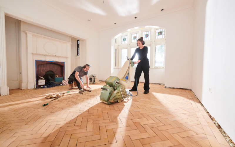 Two people work on renovating a bright, empty room with herringbone wood floors; one sands the floor while the other arranges wooden tiles near a fireplace, demonstrating expert Restoration Services in action.
