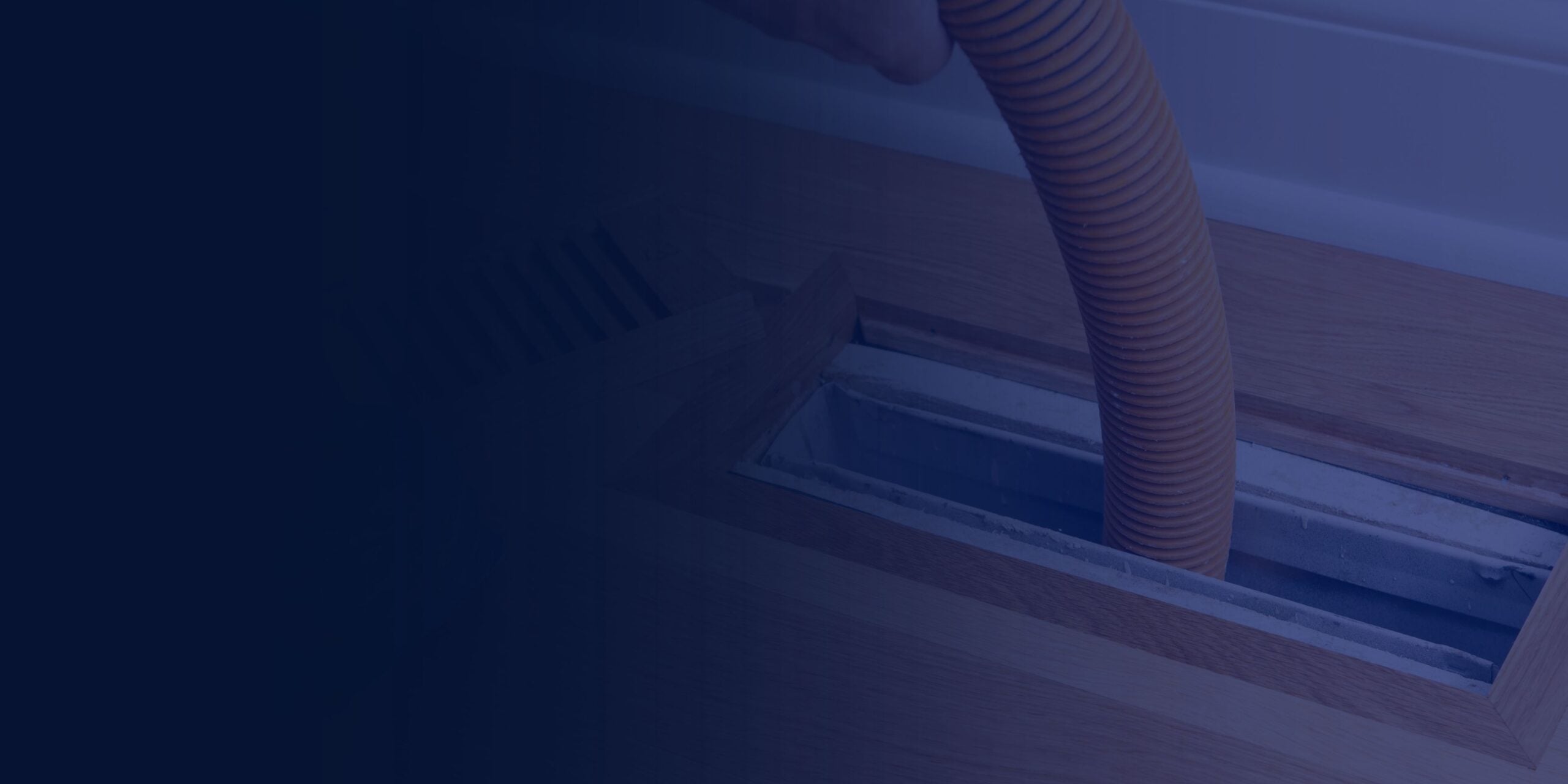 A person inserts an orange vacuum hose into a floor vent, likely to perform air vent cleaning and improve indoor air quality.