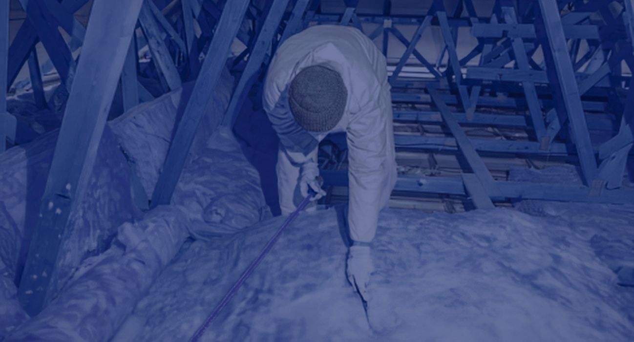 A person in protective clothing and a beanie handles insulation removal in an attic, carefully working among the surrounding wooden beams.