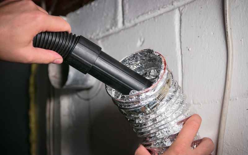 A person uses a vacuum hose to clean the inside of a flexible dryer vent duct attached to a wall, exemplifying thorough air vent cleaning.