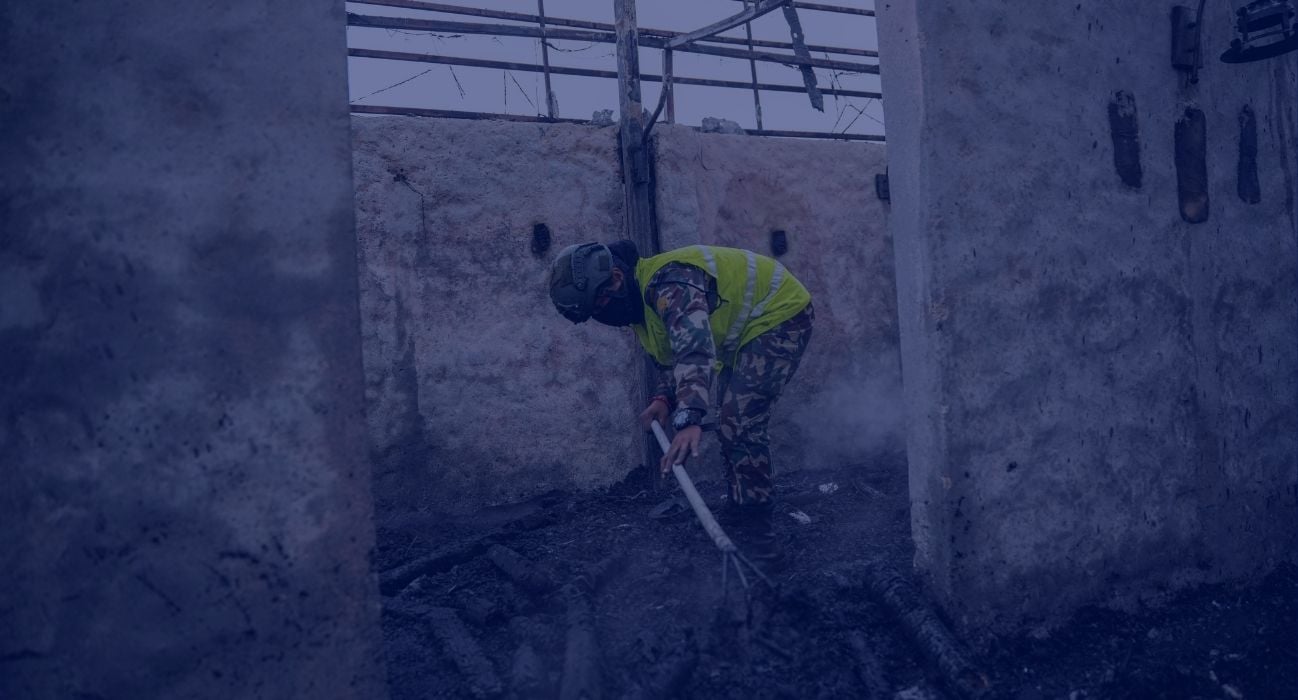 Person in safety vest and helmet sprays water onto smoldering debris and rising smoke inside a concrete structure damaged by fire.