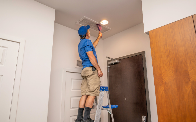 A person wearing a blue shirt and cap stands on a ladder, performing air vent cleaning on the ceiling with a pink brush inside a room with white walls and a wooden door.