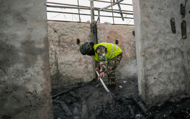 A person in camouflaged pants and a safety vest sprays water on burnt debris inside a damaged, smoky building as part of restoration services.