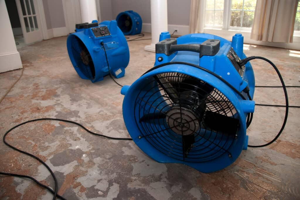 Three industrial blue air movers are placed on a bare, damaged floor in a room with columns and windows, likely for drying water damage.