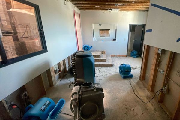 A room under renovation with exposed walls, bare ceiling beams, and several blue air movers and a dehumidifier on the floor, showcasing an active water damage restoration process.