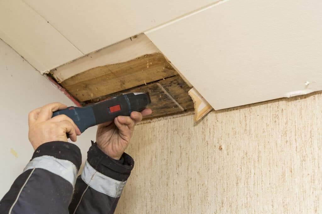A person uses a power tool to cut into a damaged section of a ceiling, exposing wooden beams and insulation.