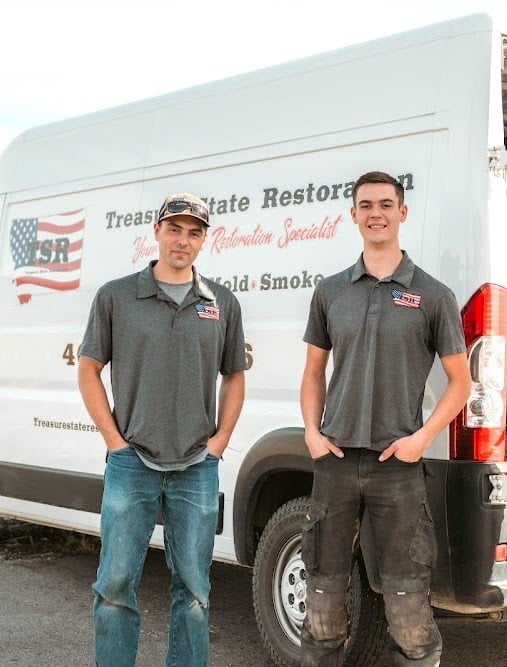 Two men in gray polo shirts stand in front of a white restoration service van with company branding and an American flag logo.
