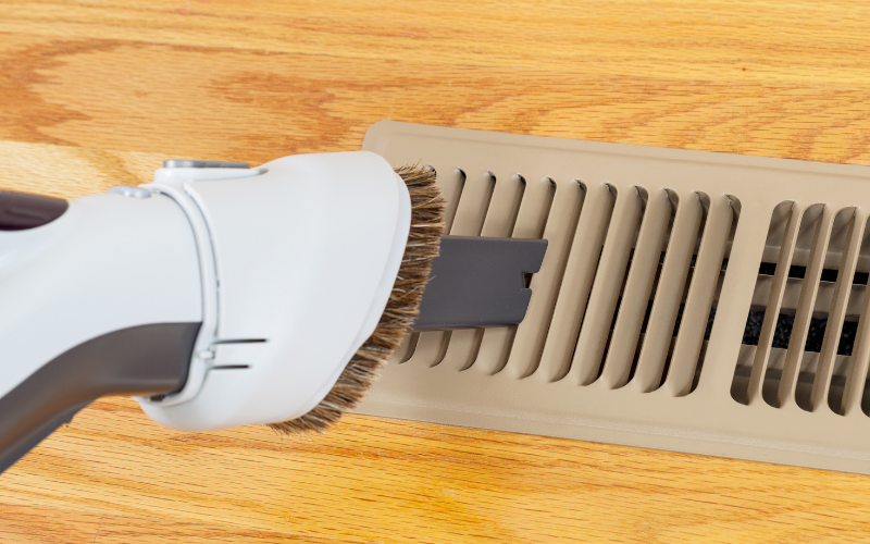 A vacuum cleaner with a brush attachment is being used to clean a floor vent on a wood floor, demonstrating effective air vent cleaning techniques for maintaining indoor air quality.