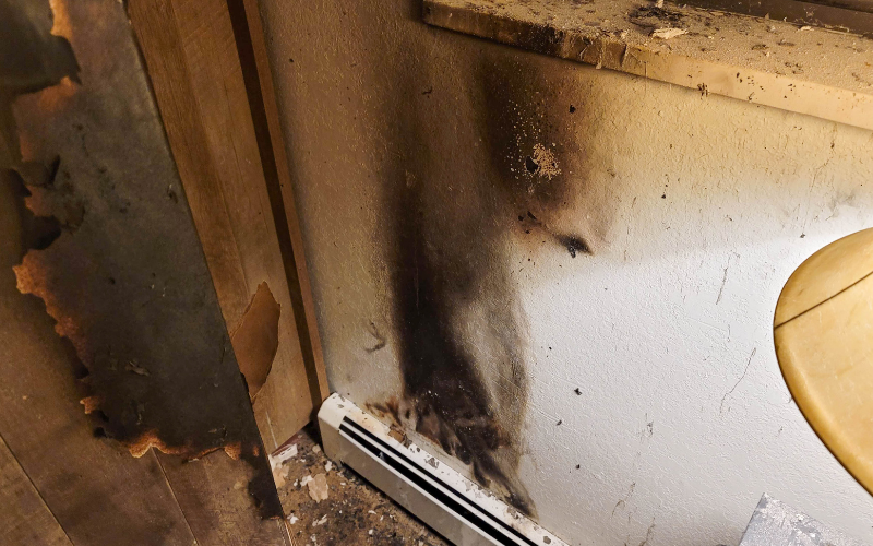 Burn marks and soot cover the wall and baseboard heater in a corner of a room, with peeling paint and scorch damage visible on adjacent surfaces—a scene in urgent need of professional restoration services.