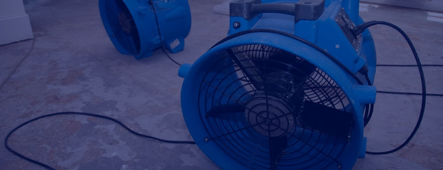 Two blue industrial air movers are placed on a concrete floor, with power cords visible, likely being used for drying or ventilation in an indoor space as part of a water damage restoration process.