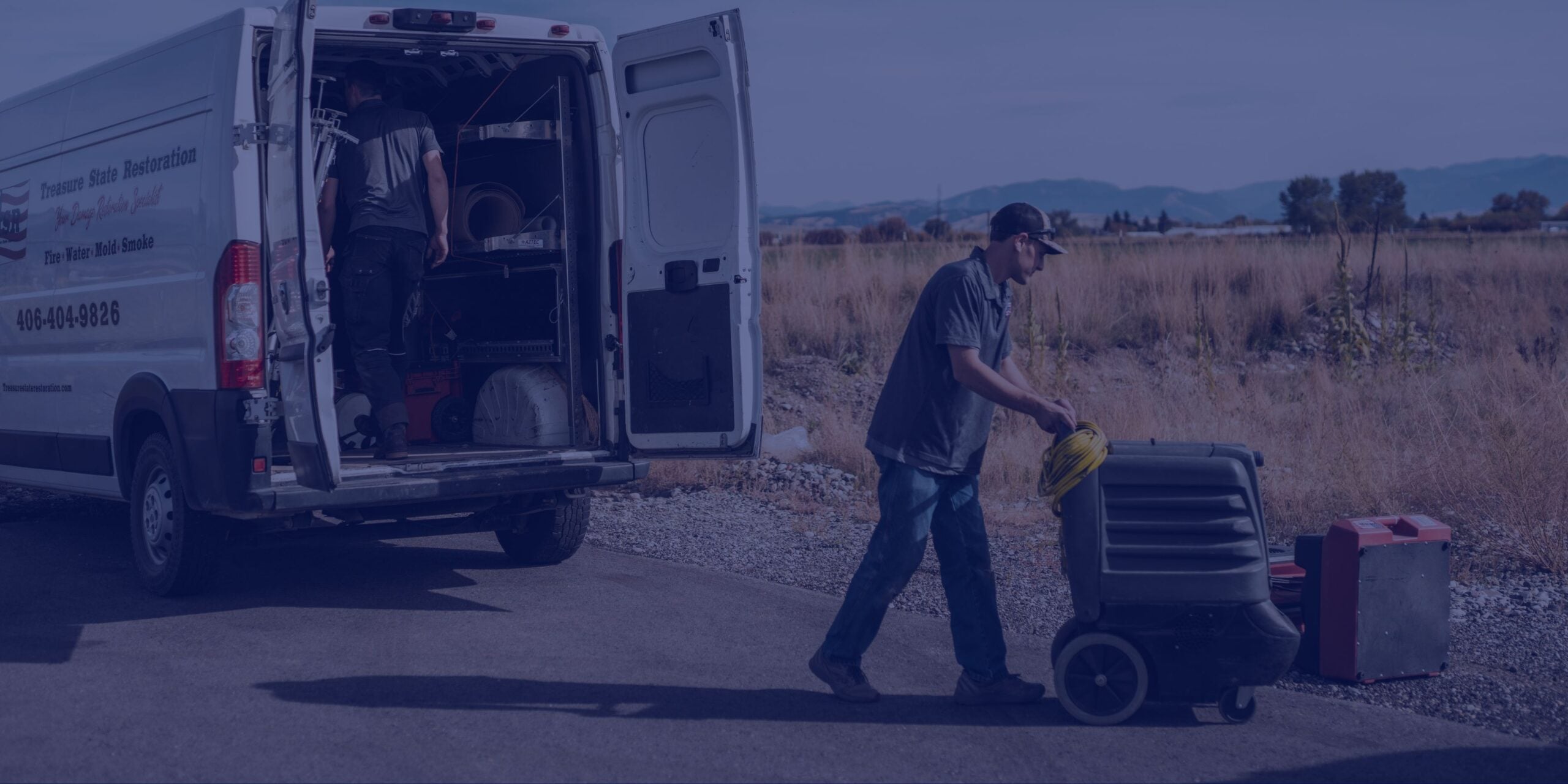 Two workers unload equipment from a white service van parked on a roadside near an open field, with one man pushing a large machine on wheels—likely used for water damage restoration.