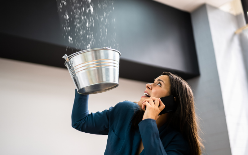 A woman holds a metal bucket under a leaking ceiling while talking on the phone, catching water as it drips down—likely calling for prompt repair services to address the urgent situation.