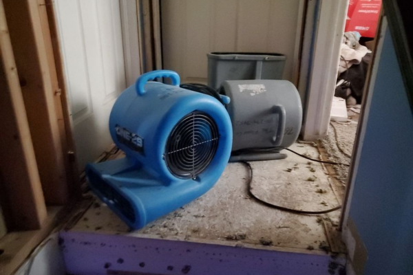 Two industrial air movers are placed on a dusty floor near an open doorway, likely for drying purposes during a water restoration project or flood cleanup.