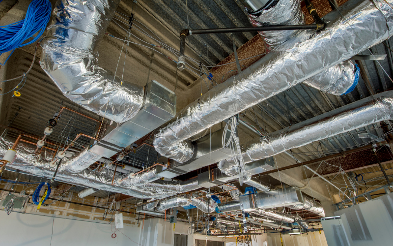 Exposed HVAC ductwork, pipes, and wiring are visible on the unfinished ceiling of a commercial building under construction or undergoing insulation removal and renovation.