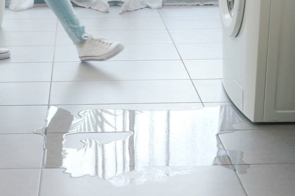 A puddle of water is on the tiled floor next to a washing machine; a person in white sneakers is walking nearby.