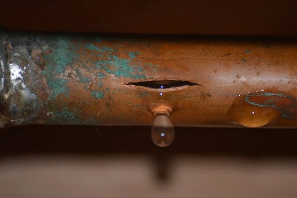 A close-up of a copper pipe with a long crack and a water droplet leaking from the damaged area.