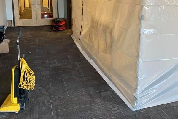 A plastic containment barrier covers part of a carpeted room. A mop bucket with a yellow cord is in the foreground, and cleaning equipment is visible near a doorway.