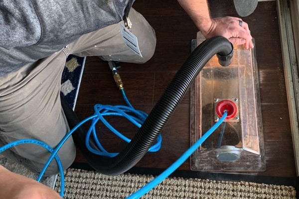 Person uses a vacuum hose attachment and blue cable to perform a cleaning or maintenance task on a hardwood floor near a rug.