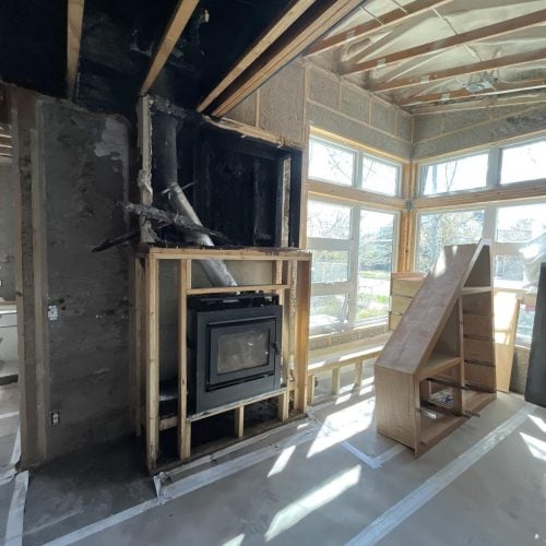 Partially constructed room with exposed framing, insulation, and a fireplace insert; large windows let in sunlight, and a wooden shelving unit is nearby—ideal for water damage restoration before finishing touches.