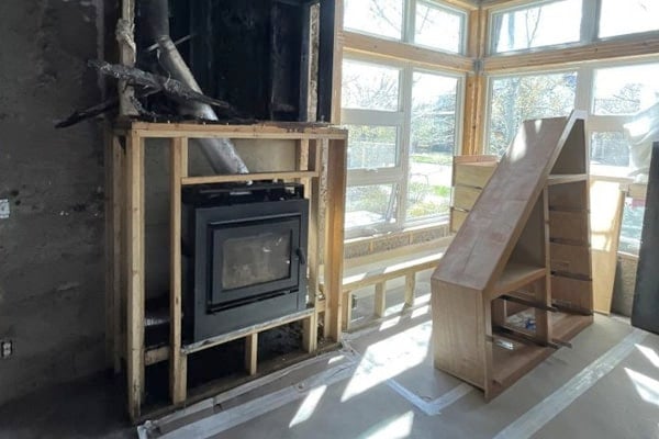 A wood stove partially installed in an unfinished room with exposed framing and a wooden staircase nearby. Sunlight streams through large windows.