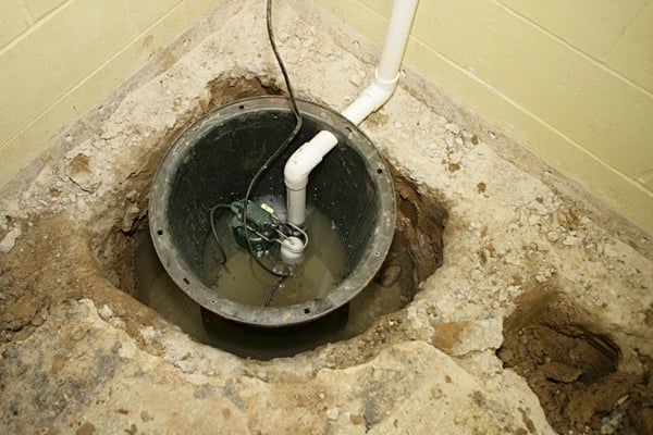 A sump pump installed in a basement floor pit, surrounded by exposed concrete and dirt, with PVC pipes attached and water inside the pit.