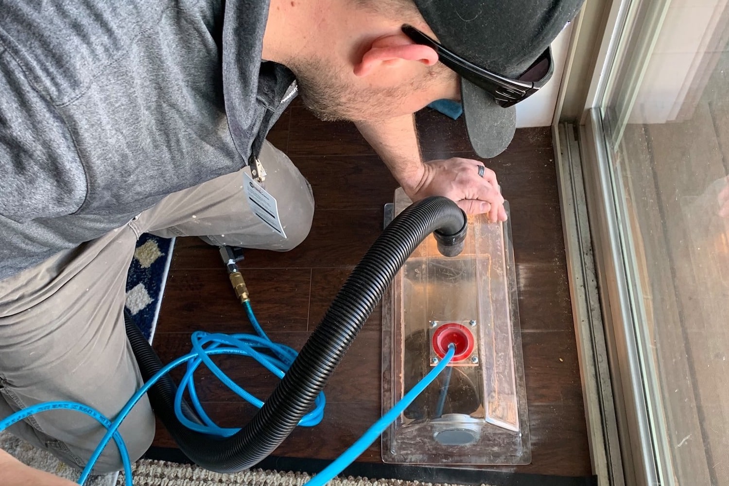 A person uses a vacuum-like machine to clean a wooden floor near a glass door, with blue and black hoses connected to the device.