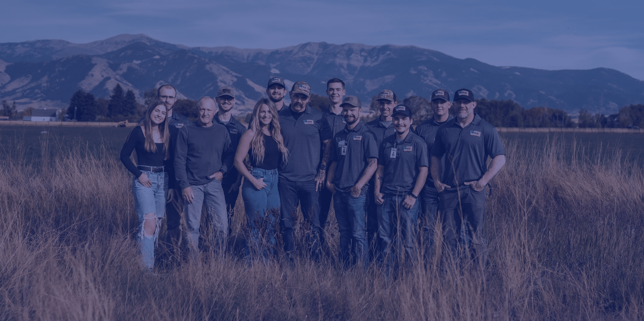 A group of thirteen people stand together in a grassy field with mountains in the background, all wearing matching shirts and caps.